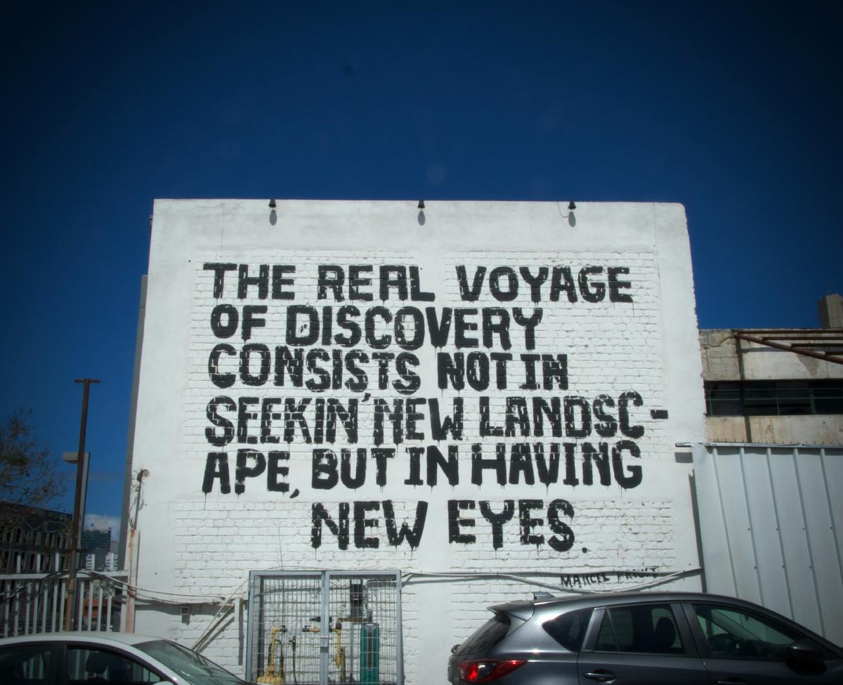 A painted wall mural reading 'The real voyage of discovery consists not in seeking new landscapes, but in having new eyes' — Marcel Proust.